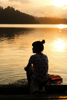 Silhouetted Thai girl is sitting and smiling on a wood raft with background of dam and sunrise at Suratthani Province of Thailand