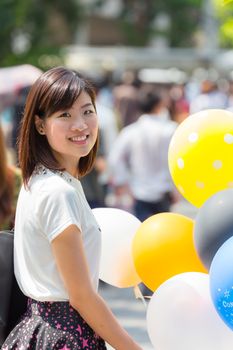 Thai girl in casual dress is smiling and holding colorful balloons in outdoor crowded place