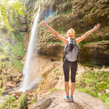 Female hiker raising arms inhaling fresh air, feeling relaxed and free in beautiful natural environment under Pericnik waterfall in Vrata Valley in Triglav National Park in Julian Alps, Slovenia.