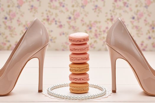 Still life. Woman essentials fashion accessories. Macarons french dessert, pearl necklace, luxury beige shoes high heels. Creative wedding set, vanilla wood, floral background. Romantic, retro vintage 