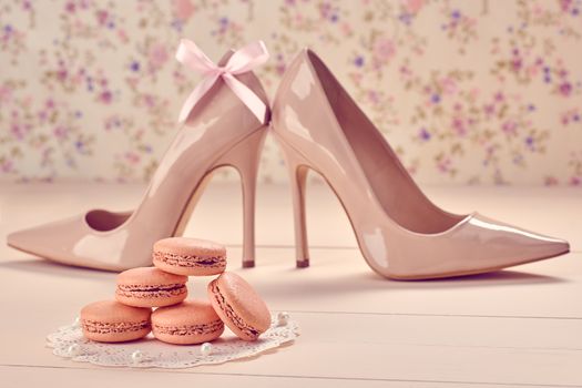 Still life. Woman essentials fashion accessories. Macarons french dessert, luxury beige shoes high heels, pearl, bow. Creative wedding set, vanilla wood, floral background. Romantic, retro vintage 