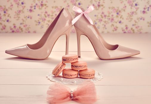 Still life. Woman essentials fashion accessories. Macarons french dessert, luxury beige shoes high heels, bow. Creative glamor wedding set, vanilla wood, floral background. Romantic, retro vintage 