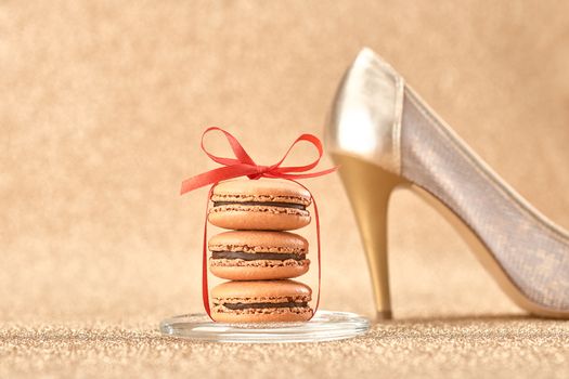 Macarons french dessert. Still life. Luxury shiny shoes high heels, red ribbon. Vintage retro romantic style. Unusual creative art greeting card, gold background, bokeh