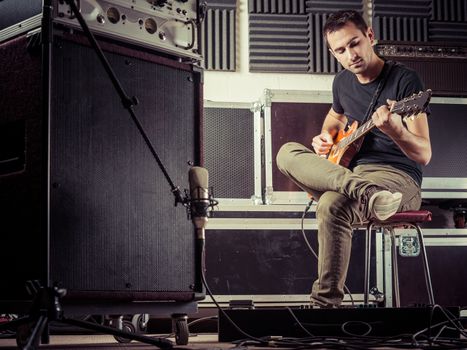 Photo of a man in his late 20's sitting in a recording studio recording his guitar tracks.