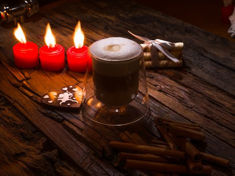 Frothy, layered cappuccino in a clear glass mug. Three red candles, cinnamon sticks and sweets. Romantic concept