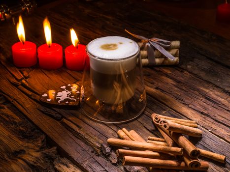 Frothy, layered cappuccino in a clear glass mug. Three red candles, cinnamon sticks and sweets. Romantic concept