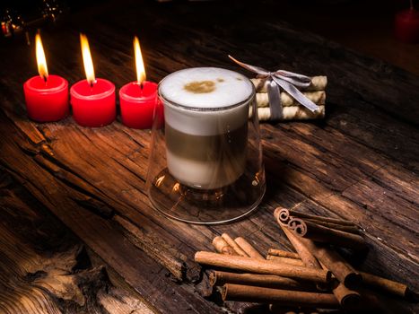 Frothy, layered cappuccino in a clear glass mug. Three red candles, cinnamon sticks and sweets. Romantic concept