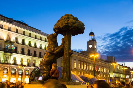 Statue of bear and madrone tree, famous symbol of city of Madrid on busy Puerta del Sol square at dusk, Spain. 