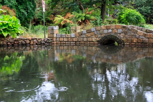 small stone arch bridge over a pond on a rainy day with trees in the background.