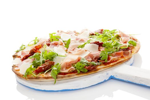 Culinary pizza with prosciutto, dry tomatoes and fresh herbs on wooden cutting board on white table. 