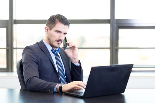 Portrait of successful corporate businessman in bright modern office focused on data on his laptop computer while talking on mobile phone. Business and entrepreneurship concept.