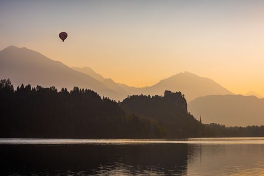 Silhouette of Bled Castle on a Lake at Sunrise with Mountains and Hot Air Balloon Flying