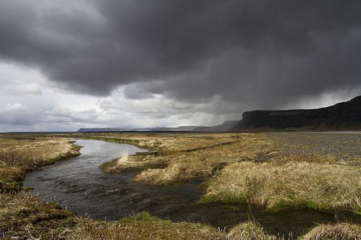 Horizontal panorama view of an Icelandic river running through the grass with rain stormy clouds above and mountains on the right side