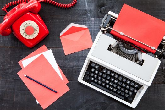 The image represents a retro typewriter , a vintage red telephone and some coloured paper on a dark wood background with a rede paper conceptualizing this colour like danger, love or something different