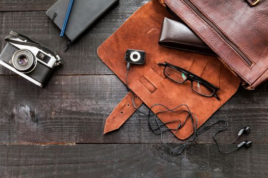 The image shows a retro or hipster bag for man or woman with stuff such as glasses , vintage camera, leather bag and a book note on a wooden background