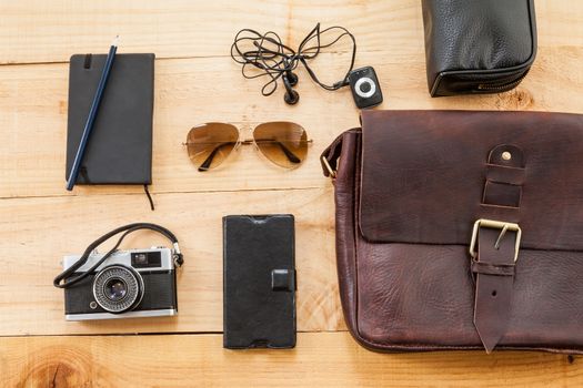 The image shows a retro or hipster bag for man or woman with stuff such as glasses , vintage camera, leather bag and a book note on a wooden background