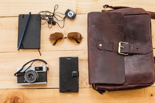 The image shows a retro or hipster bag for man or woman with stuff such as glasses , vintage camera, leather bag and a book note on a wooden background