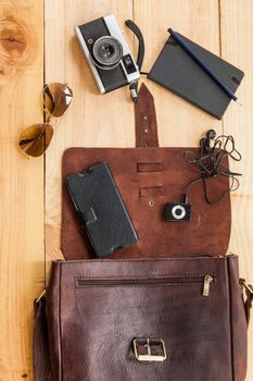 The image shows a retro or hipster bag for man or woman with stuff such as glasses , vintage camera, leather bag and a book note on a wooden background