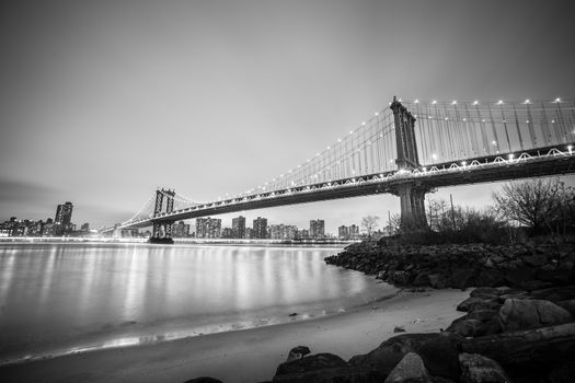 Manhattan bridge and New York City downtown skyline at dusk with skyscrapers illuminated over East River panorama. Black and white image.