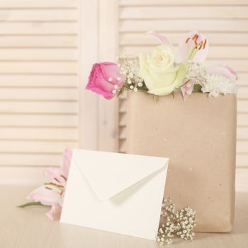 Flowers in paper bag and blank envelope on wooden background