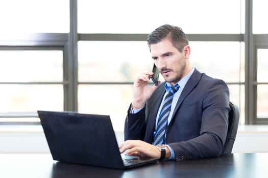 Portrait of successful corporate businessman in bright modern office focused on data on his laptop computer while talking on mobile phone. Business and entrepreneurship concept.