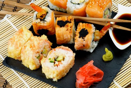 Arrangement of Various Maki Sushi with Tempura Crab, Smoked Salmon and Eel with Black Caviar on Stone Plate with Soy Sauce, Ginger and Pair of Chopsticks closeup on Straw Mat background