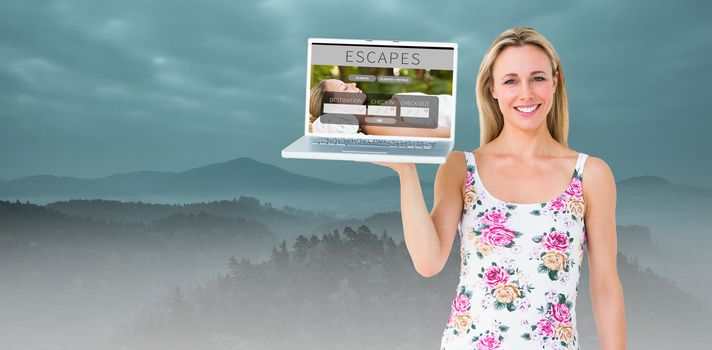 Smiling blonde holding laptop and posing against trees and mountain range against cloudscape