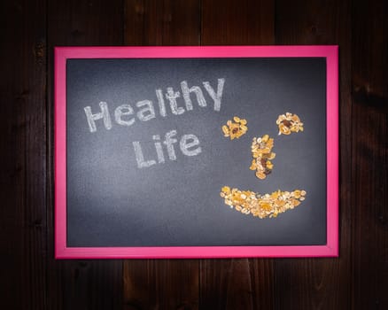 In the picture a blackboard, on the left side with written "Healthy Life" and on the right side a mixture of cereals which draw a smile on wooden background