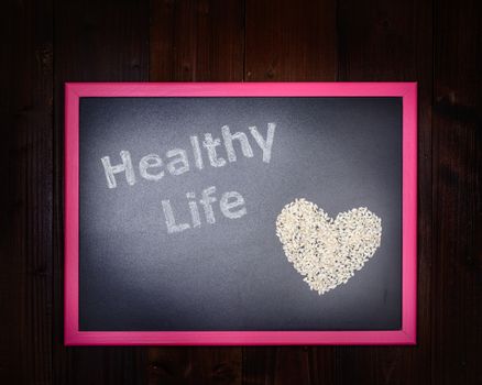 In the picture a blackboard, on the left side with written "Healthy Life" and on the right side a rice grains which draw a heart on wooden background