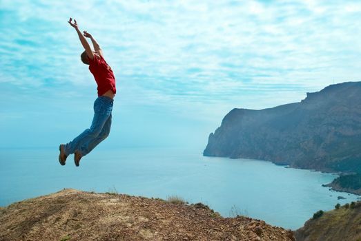 Man jumping cliff against sea and mountain with blue sky