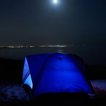 Blue illuminated tent at night on the beach. Moon, stars and reflection in water