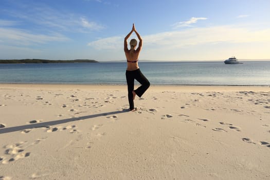 Woman balanced in a yoga pose standing barefoot on one leg with her hands joined above her head at the beach in late afternoon sunlight, drawing long shadows on the sand. Asana vrksasana.