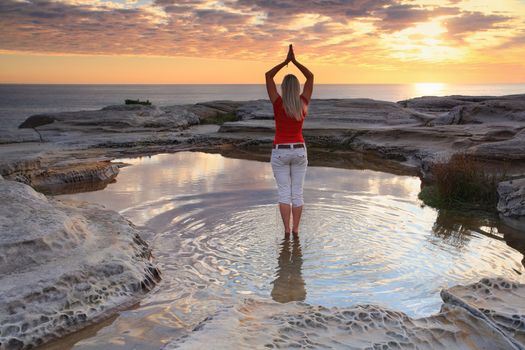 A woman standing by the ocean at sunrise, practicing yoga, meditation, pose.  Rejuvenating the soul, quiet time and solitude in natures beautiful surrounds.