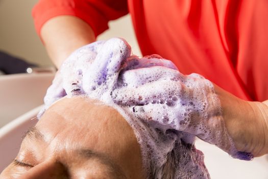 Close up of face of man is getting a hairwash by a hairdresser, with relaxation. The beautician is making foam from shampoo
