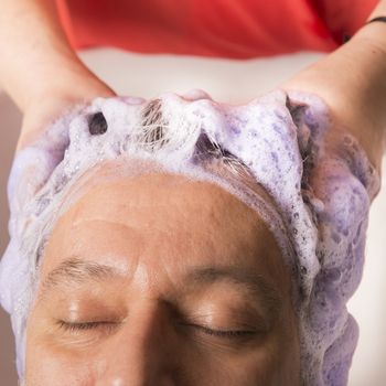 Close up of face of man is getting a hairwash by a hairdresser, with relaxation. The beautician is making foam from shampoo