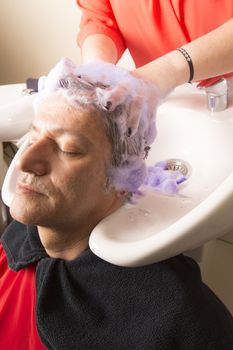 Close up of face of man is getting a hairwash by a hairdresser, with relaxation. The beautician is making foam from shampoo