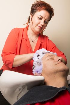 Close up of face of man is getting a hairwash by a hairdresser, with relaxation. The beautician is making foam from shampoo