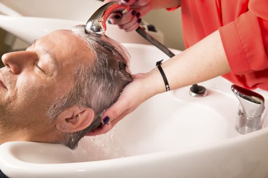 Close up of face of man is getting a hairwash by a hairdresser, with relaxation. The beautician is making foam from shampoo