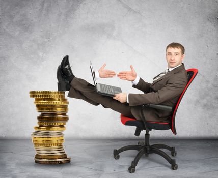 Businessman giving hand to laptop with pile of coins on grey wall background