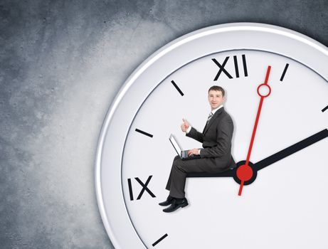 Businessman sitting on clockhand and working on laptop on grey wall backgound