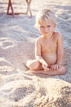 Little boy with blue eyes and white hair sitting on white sand and look up at the camera