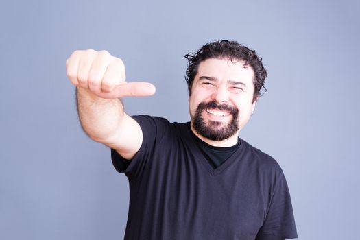 Waist Up Portrait of Joyful Laughing Man with Beard and Curly Hair Giving Side Thumb Hand Gesture Towards Camera in Studio with Gray Background and Copyspace