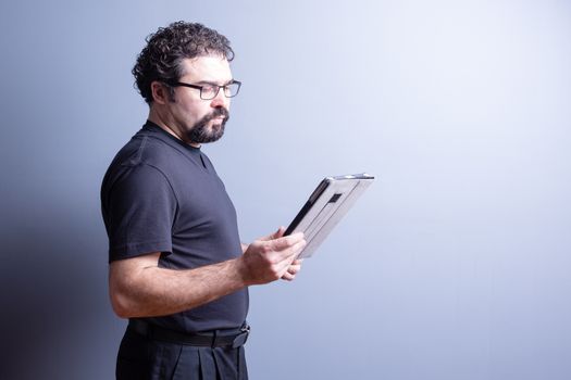 Profile of Man Wearing T-Shirt and Eyeglasses Looking Serious and Reading from Computer Tablet in Studio with Gray Background, Side Lighting and Copy Space