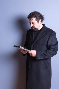 Three Quarter Length of Man with Beard and Curly Hair Wearing Long Dark Trench Coat and Looking Down Seriously at Computer Tablet in Studio with Gray Background and Copy Space