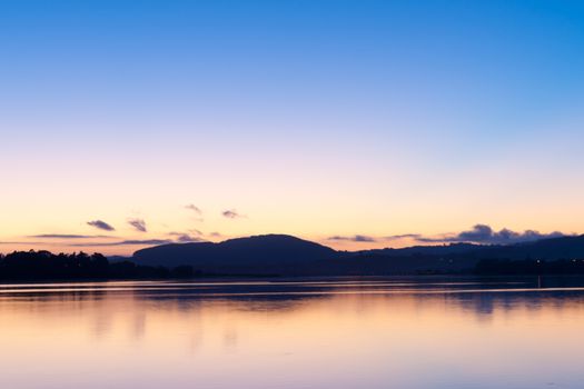 Sunrise over the bay blue sky transitions to pink and orange from above the horizon and over calm water.