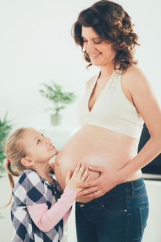 Cute smiling young girl cuddling pregnant belly of her pregnant mother.