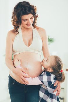 Cute smiling young girl cuddling pregnant belly of her pregnant mother.