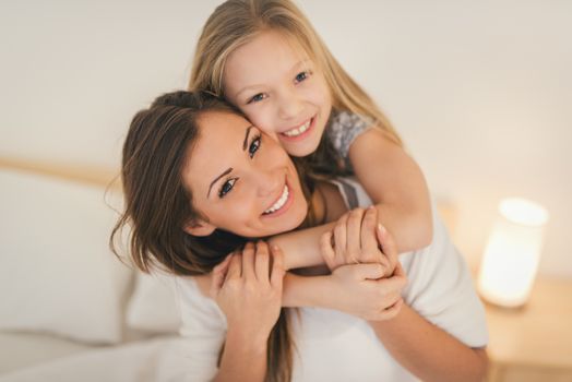Beautiful smiling mother and her daughter hugging in bed. They are sitting on bed at pajamas and smiling looking at camera.