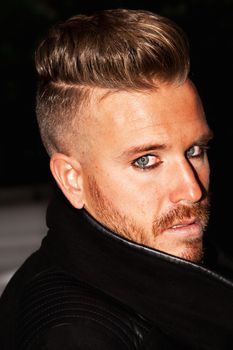 Portrait of a fashionable young man with blue eyes, stylish haircut and posing with a black jacket. Close up. Vertical image.