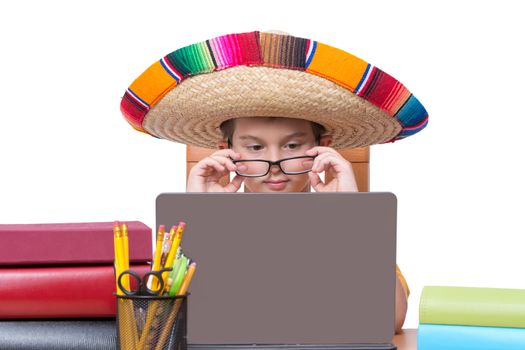 Young Boy Wearing Eyeglasses and Colorful Mexican Sombrero Hat Looking Down at Laptop Computer Screen While Sitting at Desk Surrounded by Colorful Books and Binders and Writing Supplies